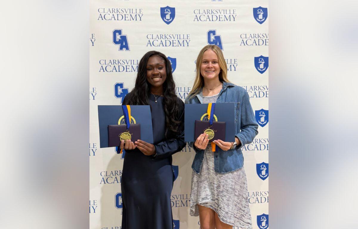 Two graduates stand side by side, smiling, holding blue folders with gold medals in front of a Clarksville Academy backdrop.