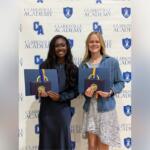 Two graduates stand side by side, smiling, holding blue folders with gold medals in front of a Clarksville Academy backdrop.