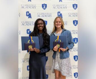 Two graduates stand side by side, smiling, holding blue folders with gold medals in front of a Clarksville Academy backdrop.