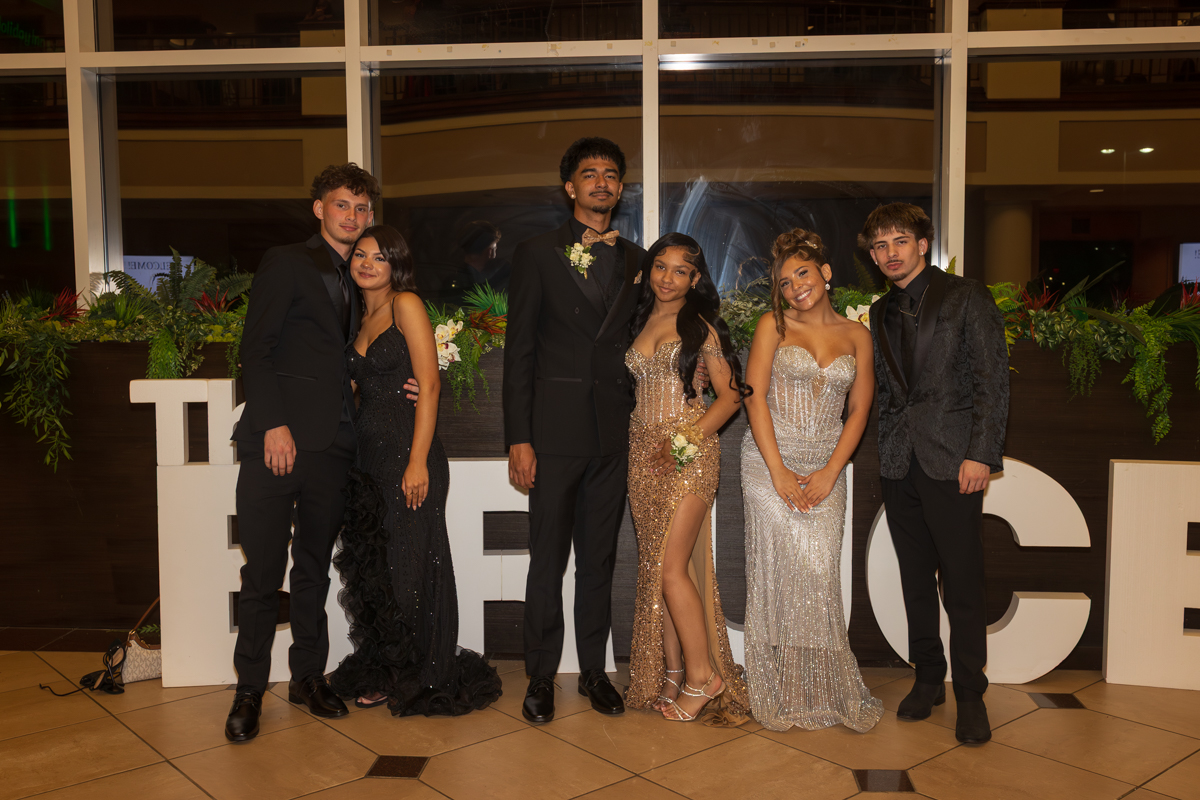 Group of six young adults in formal wear posing for a photo at a decorated event venue.