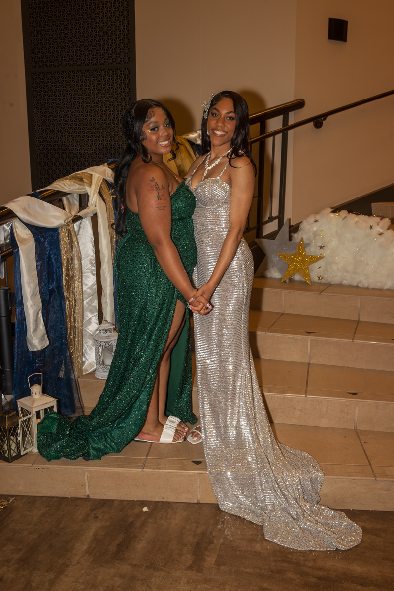 Two women in formal gowns pose together on a stair landing at a festive event, smiling at the camera; one in a green sequined dress, the other in a silver glitter gown holding hands.