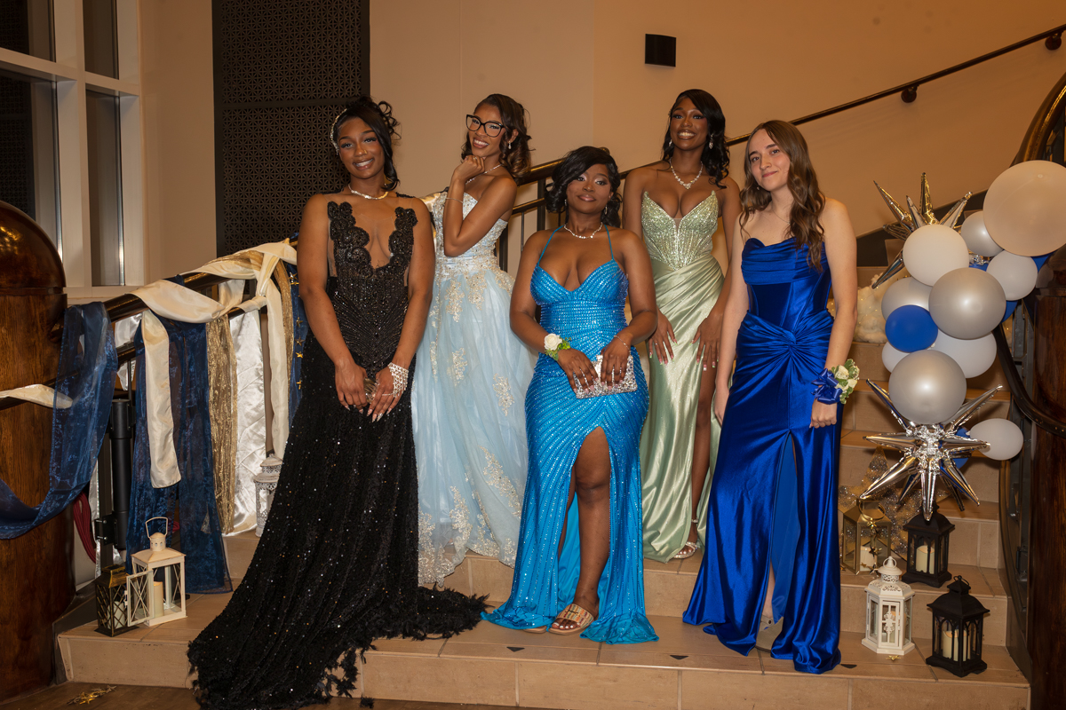 Five young women in formal evening gowns pose on a stairway with blue, black, and silver decor.
