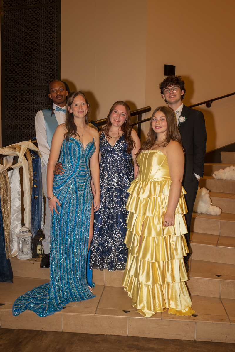 Five teens in formal wear posing on a staircase at a prom, with two boys in suits and three girls in colorful gowns.