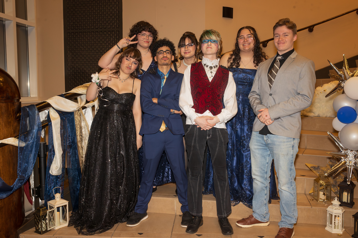 Seven teens in formal wear pose on a staircase at a prom, wearing blue gowns, a blue suit, and a red-vested outfit in center.