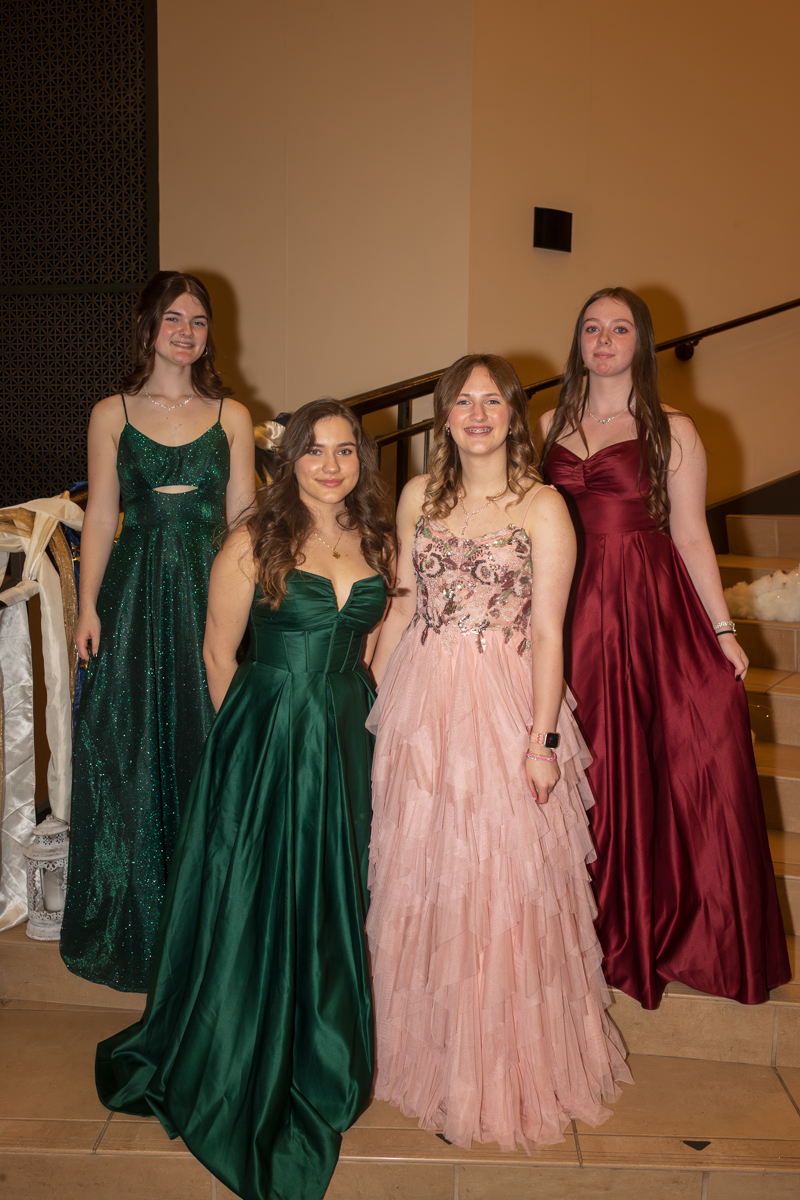 Four young women in formal gowns pose on a staircase at a formal event indoors, wearing green, emerald, pink, and burgundy dresses.