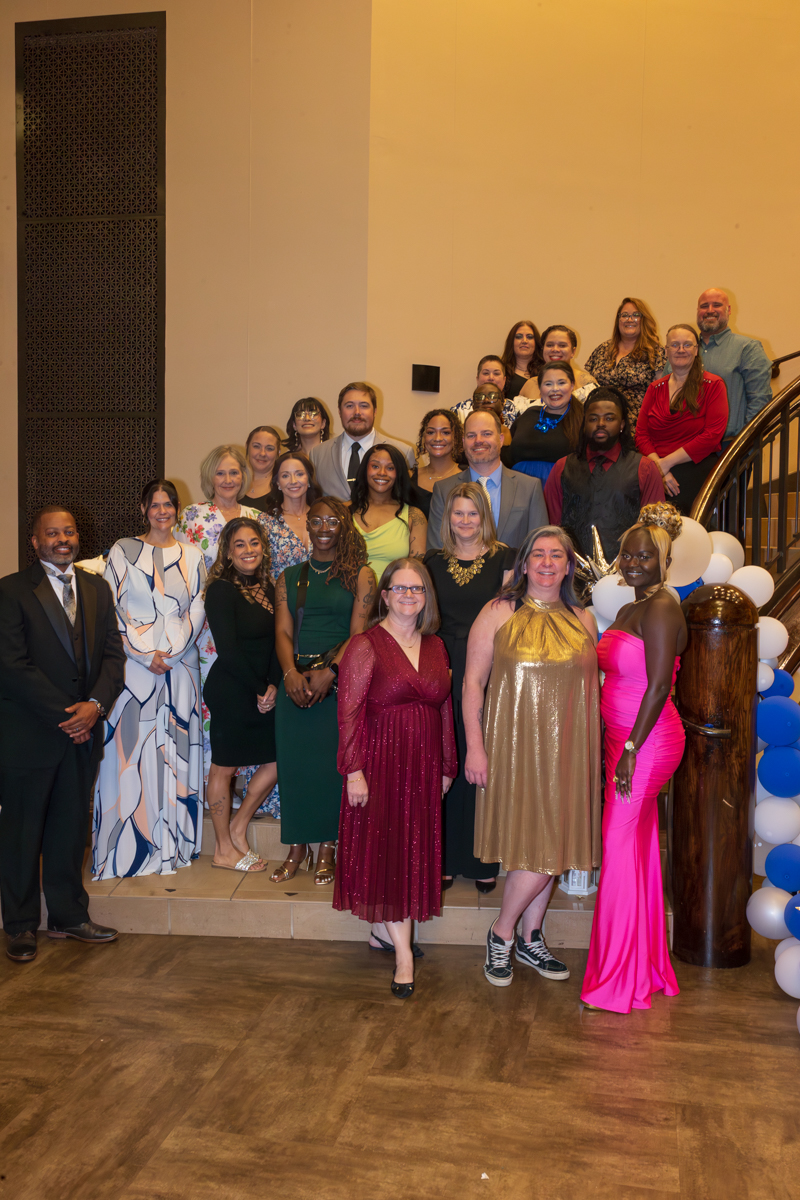 Group of men and women in formal attire posing for a group photo on a staircase at a celebration, with blue and white balloons nearby.