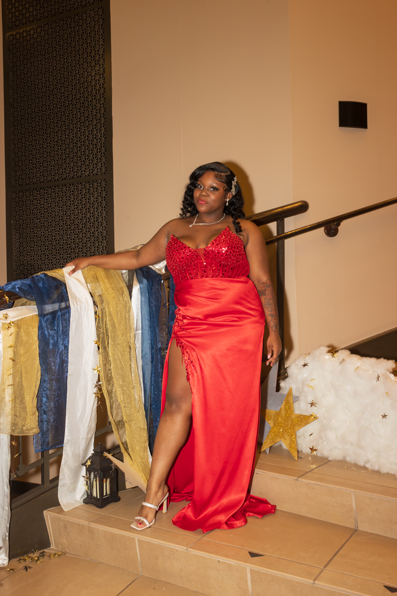 Woman in a red sequined evening gown with a high slit posing on a decorated staircase, with blue, gold, and white fabric draped along the railing and star decor nearby.