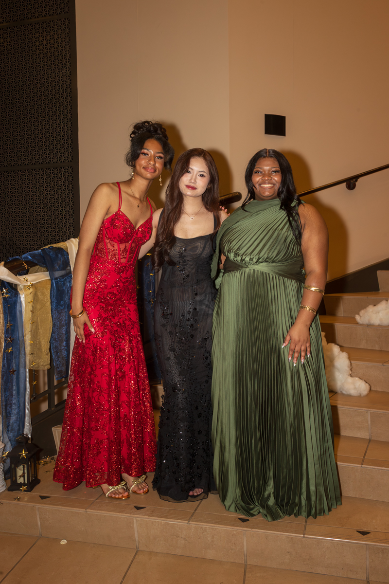 Three women in formal gowns pose on a staircase at an event, wearing red, black, and green dresses and smiling nearby decorations.