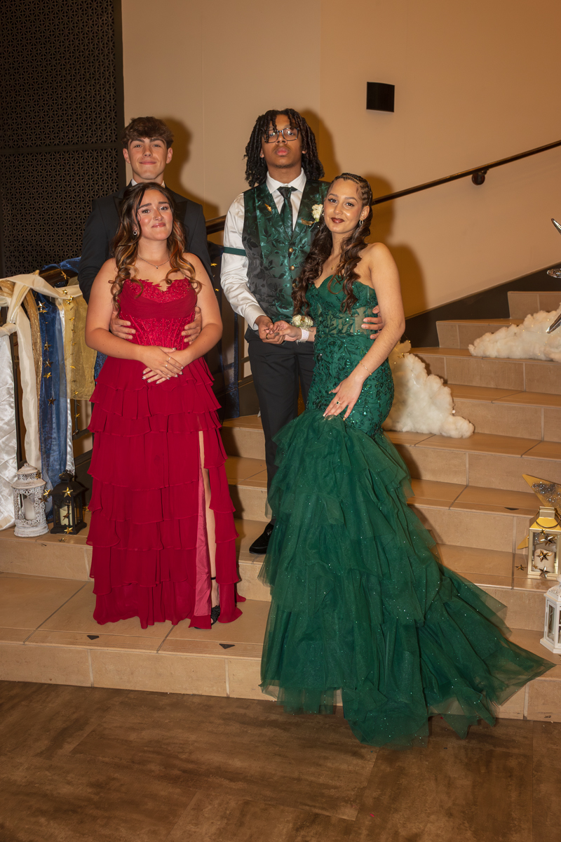 Four teens in formal wear posing on a staircase at a prom: two girls in red and green gowns and two boys in suits.