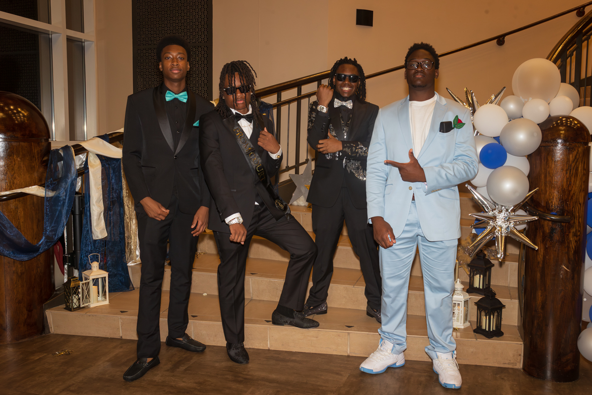 Four young men in suits pose on a staircase at a formal event, with blue and white decorations in the background.