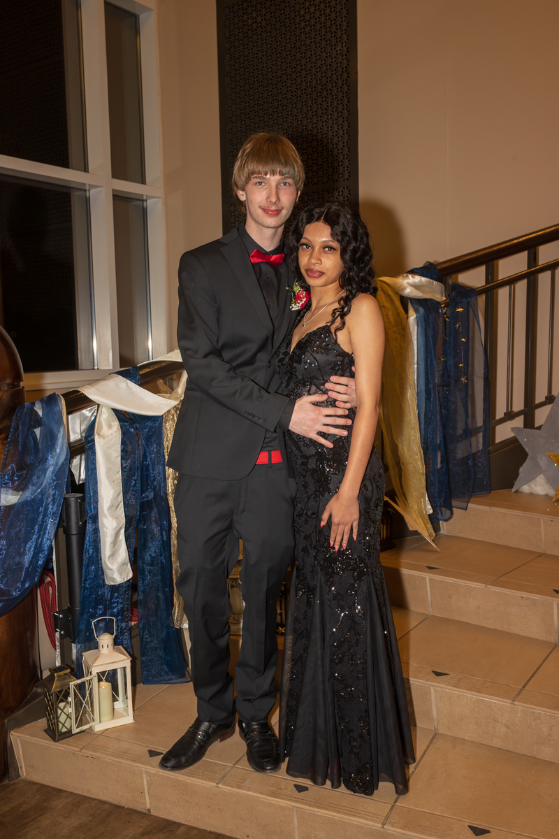 Young couple in formal wear posing on a staircase; man in a black suit with red bow tie and boutonniere, woman in a black sequined gown beside him.