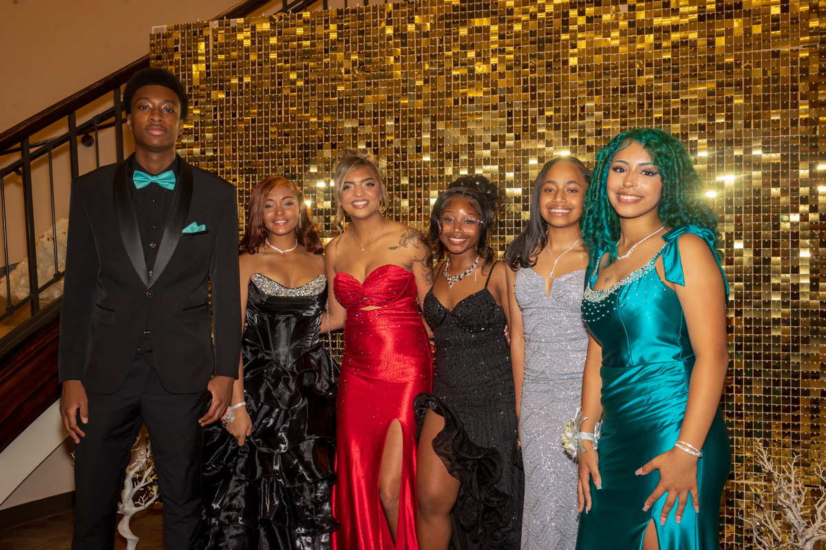 Six teens in formal attire posing together at a gala, smiling in front of a gold mosaic backdrop.
