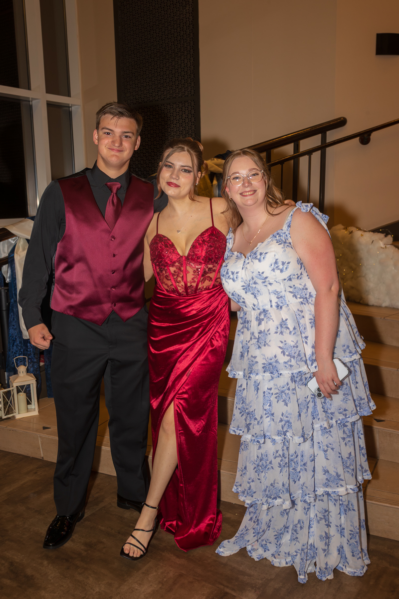 Three friends posing together at a formal event, the woman in the middle wearing a red velvet gown with a thigh-high slit, flanked by a man in a burgundy vest and a woman in a blue floral dress holding a phone.
