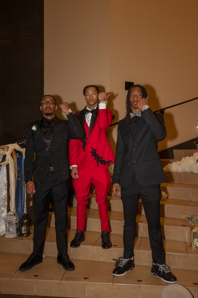 Three men in formal suits pose on a staircase at a formal event; the center man wears a red suit with black embroidery and bow tie, flanked by two men in black suits with bow ties, all making confident gestures toward the camera.