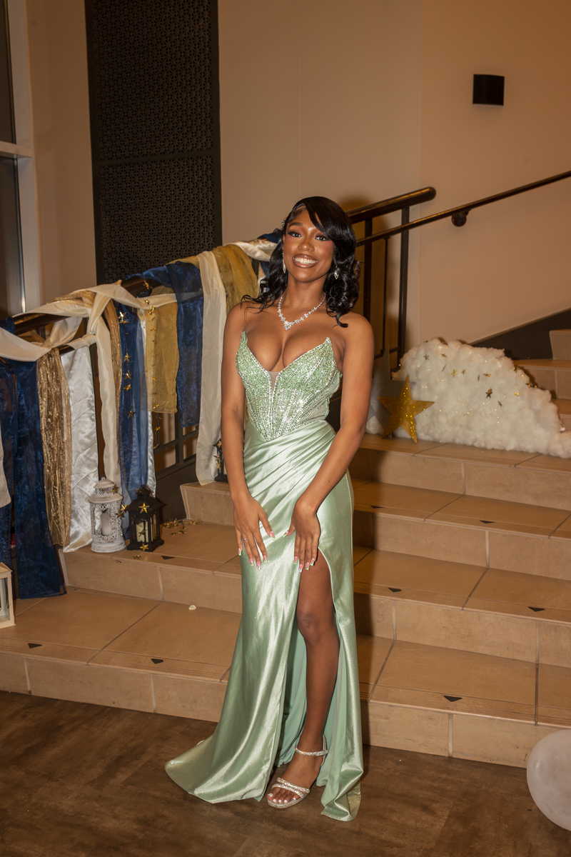 Smiling woman in a mint-green, sequined evening gown with a thigh-high slit, posing on a decorated indoor staircase.
