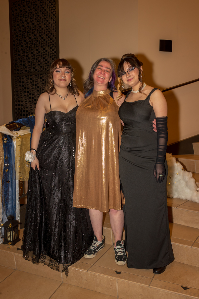 Three women in formal gowns pose together on a staircase at an evening event. Left: black sequined dress, center: metallic gold dress with sneakers, right: black gown with gloves and glasses.