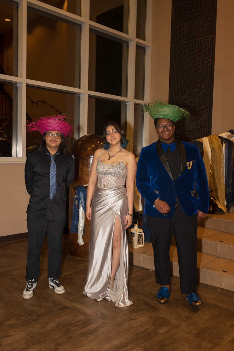 Three young adults posing indoors at a formal event: a woman in a silver gown between two men in suits with colorful hats.