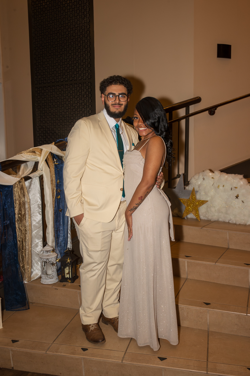 Couple dressed formally posing on a staircase with festive decor and stars in the background.