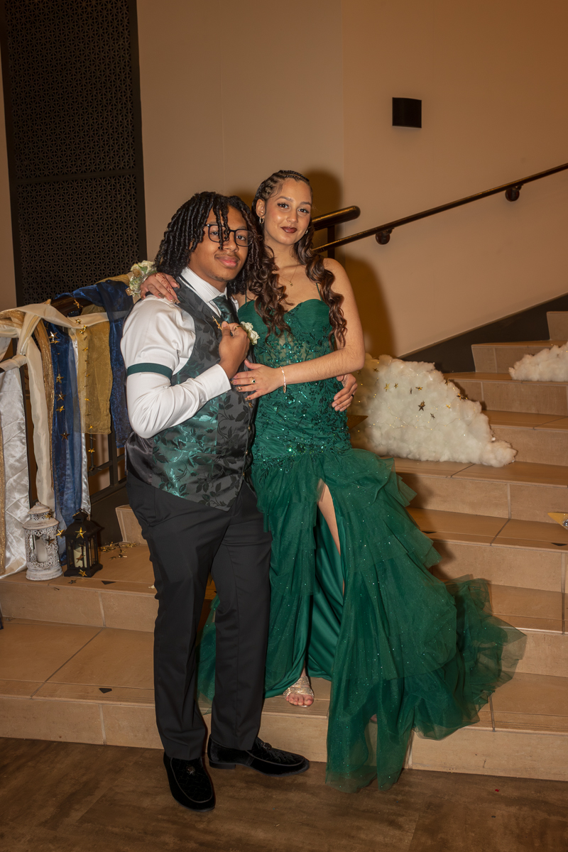 Two young people pose together on a staircase at a formal event; girl in an emerald green gown with a high slit and tiara, boy in a vest and dress shirt holding her arm.