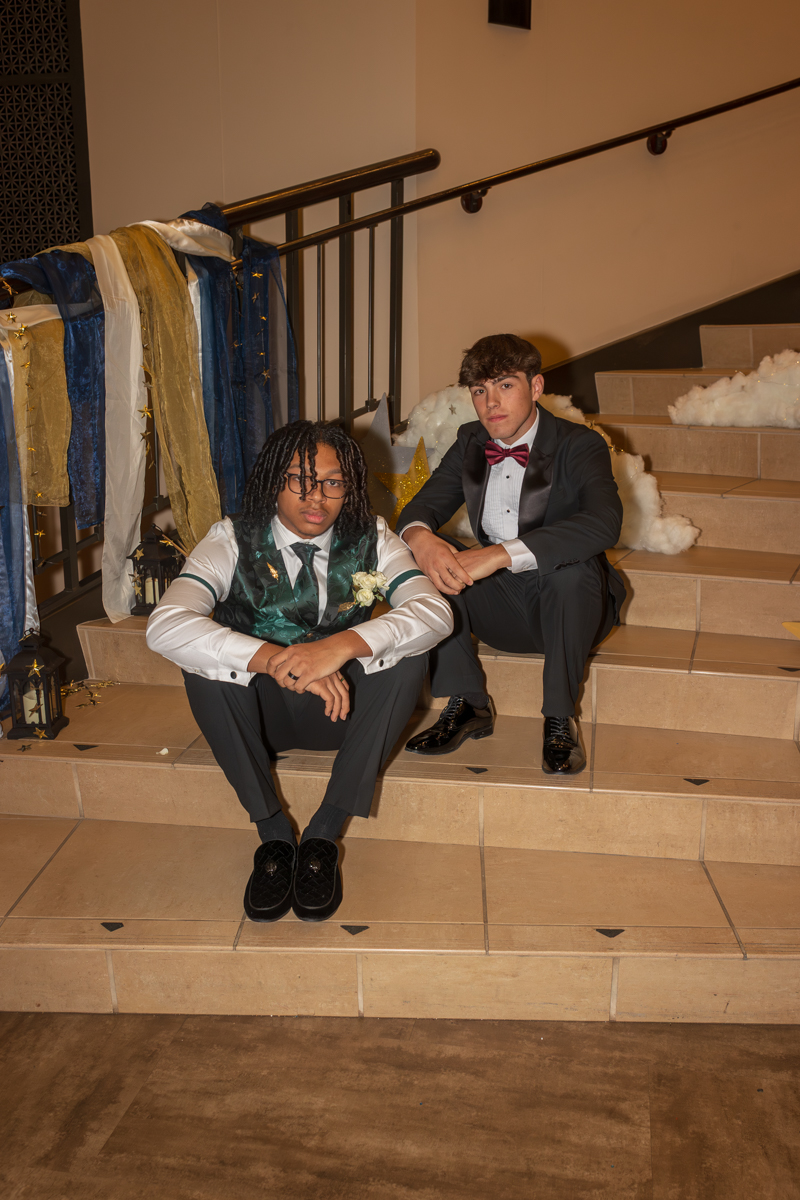 Two young men in tuxedos sit on a decorated staircase, draped in blue, white, and gold fabric with lanterns nearby.