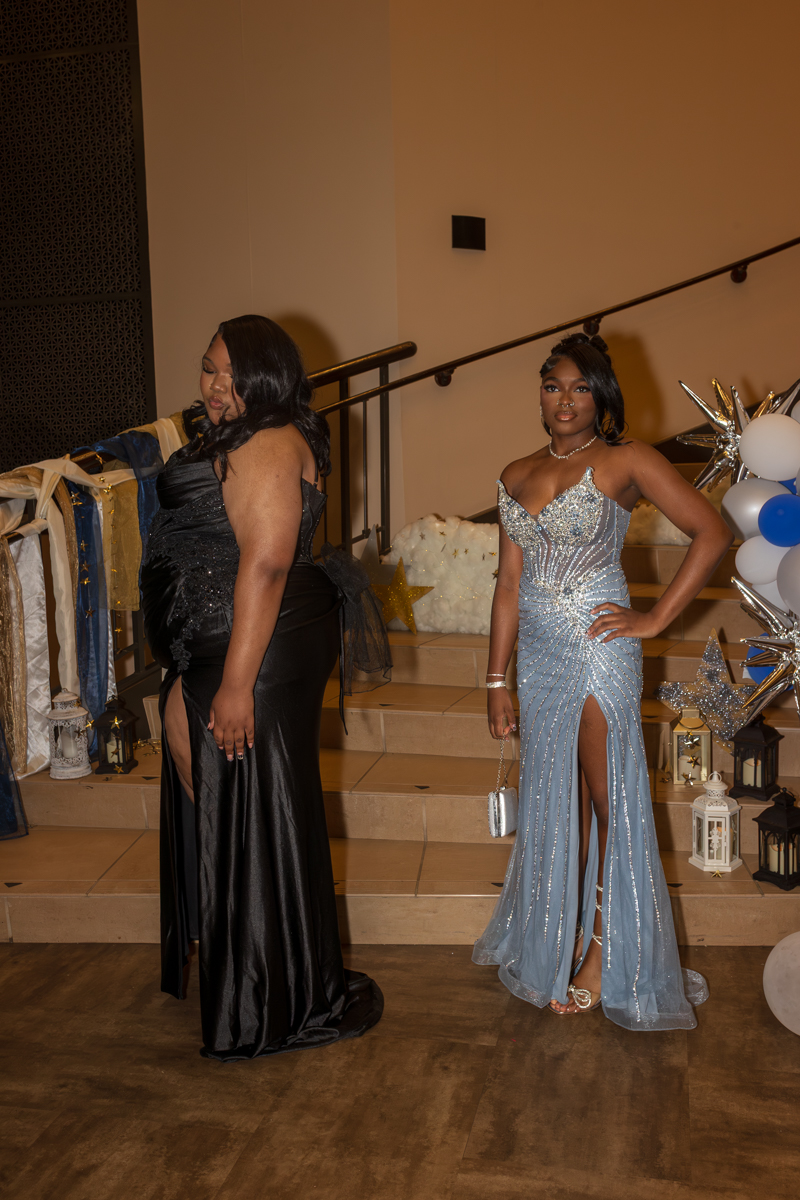 Two women in formal gowns pose on a stairway at a decorated event, one in a black dress and the other in a light blue embellished gown with a high slit.