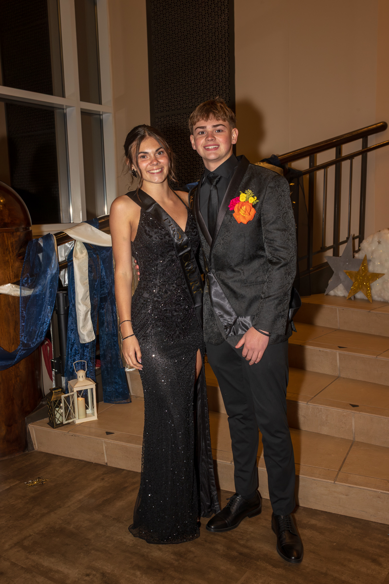 Two teens in formal attire pose on a staircase at a prom; girl in a black sequined gown, boy in a dark suit with a boutonniere.