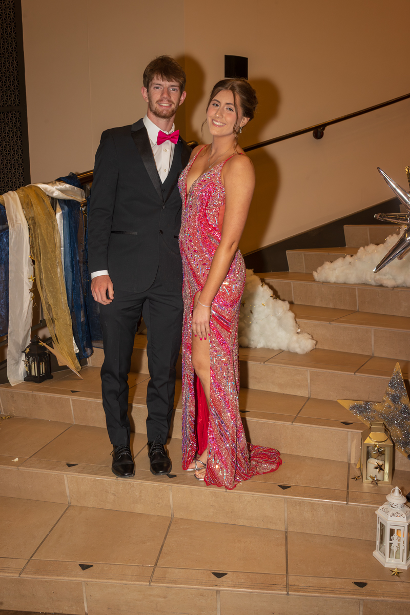 Couple in formal attire on a staircase: man in a black suit with pink bow tie and a pink sequined gown worn by a smiling woman with a thigh-high slit.