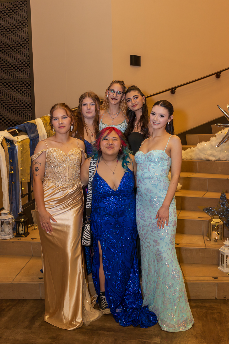 Six young women in formal gowns pose together on a staircase at a fancy event, smiling for the camera.