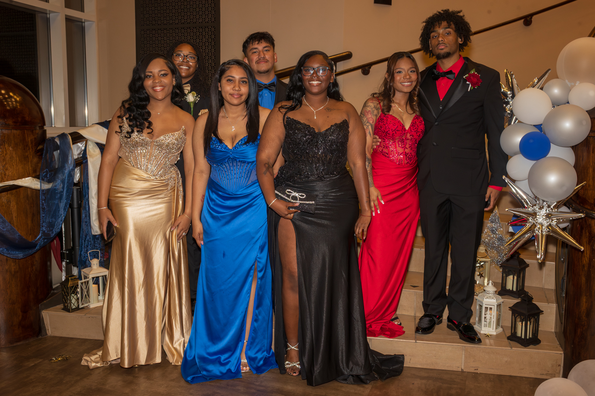Group of eight young adults in formal attire posing on a staircase with balloons and lanterns for a gala photo.