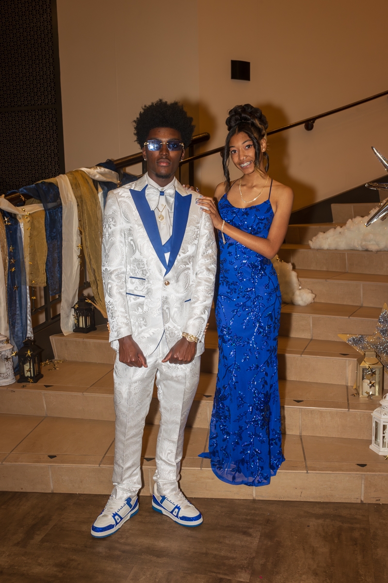 Couple posing on stairs at a formal event: man in a white suit with blue accents and matching sneakers, wearing sunglasses, beside a woman in a blue sequined gown.