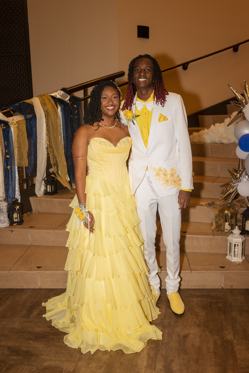 Couple posing at a formal event: woman in a yellow tiered ball gown and corset, man in a white suit with yellow accents, on a staircase backdrop.
