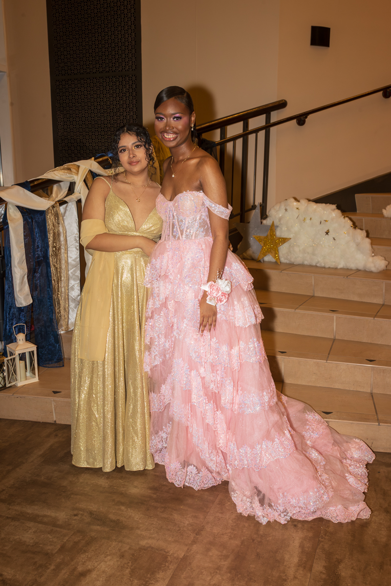 Two young women in formal gowns pose together on a stairway at a gala, one in a gold dress and the other in a pink lace ball gown with off-the-shoulder sleeves.