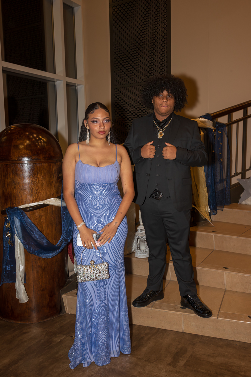 Couple in formal wear posing on a staircase; woman in a blue sequined gown with a silver clutch, man in a black suit with gold chains beside her.