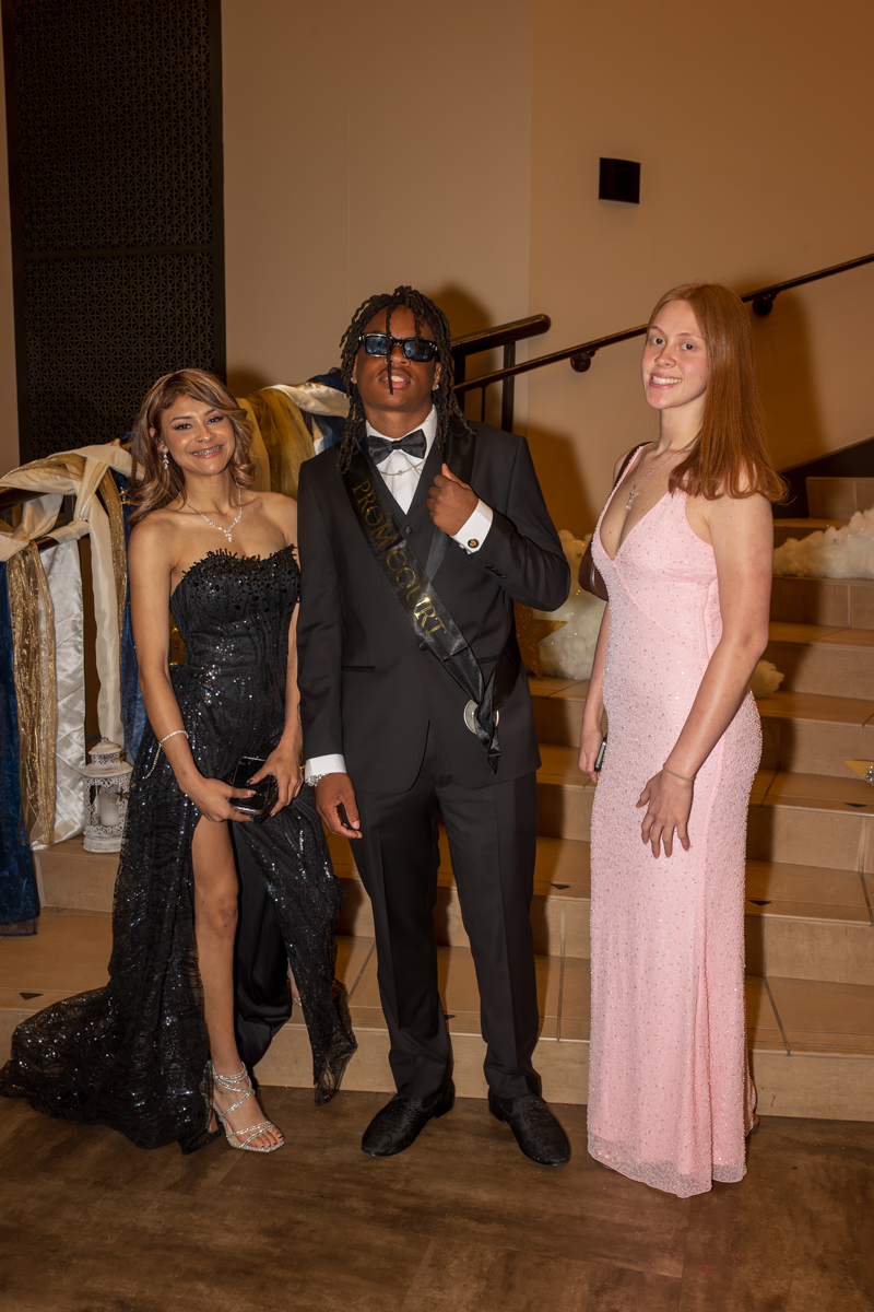Three teens in formal attire pose on a staircase at a prom event: a girl in a black sequined gown with a slit, a boy in a black tux with sunglasses and a 'PROM COURT' sash, and a girl in a pink sequined gown.
