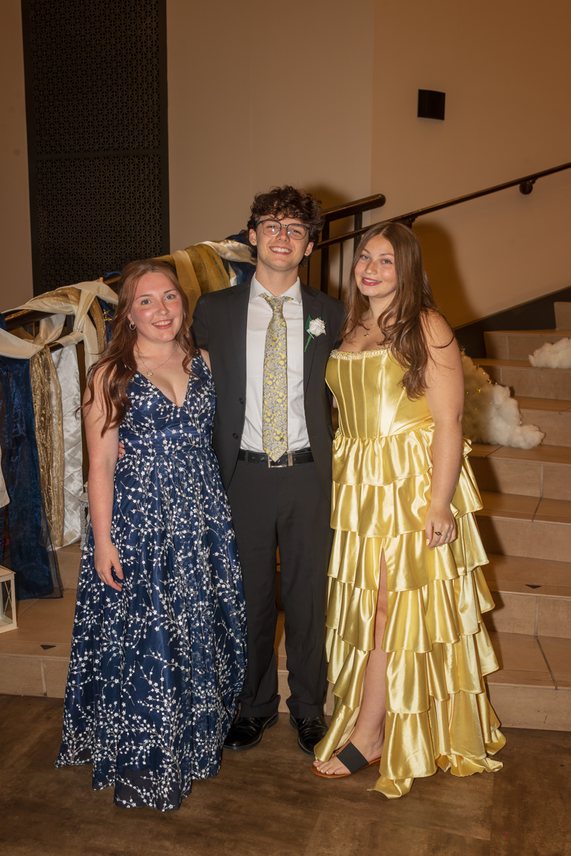 Three friends in formal attire posing together on a stairway at a prom or formal event, smiling at the camera.
