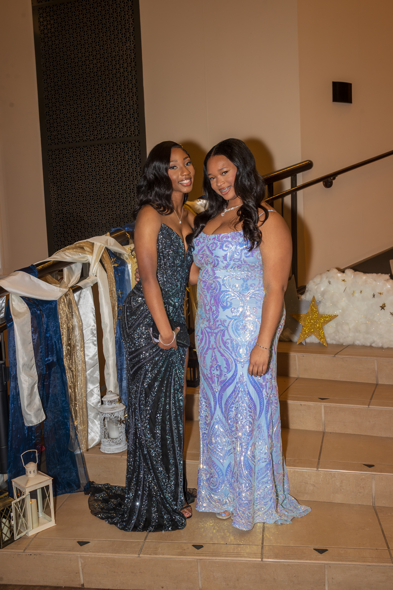 Two women in elegant evening gowns pose together on a staircase, smiling at a formal event with decorative drapery and stars nearby.