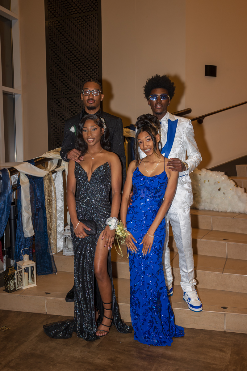Group of four teens posing on a staircase at a formal event: two women in sparkling floor-length gowns (one black, one blue) and two men in suits behind them, one in a white patterned suit with blue accents and sunglasses.