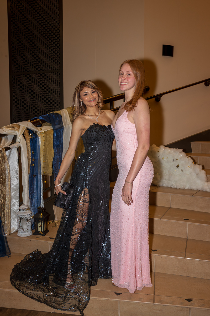 Two young women in formal gowns pose on a decorated staircase at a gala, one in a black sequined dress and the other in pink.