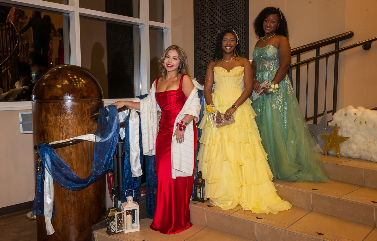 Three women in formal gowns pose on a staircase at a decorated event: red dress, yellow ballgown, and teal gown smiling at the camera.