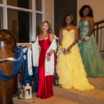 Three women in formal gowns pose on a staircase at a decorated event: red dress, yellow ballgown, and teal gown smiling at the camera.