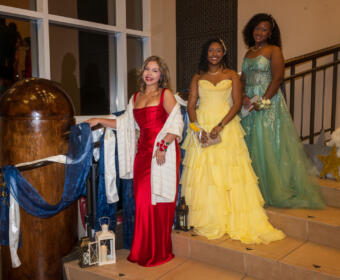 Three women in formal gowns pose on a staircase at a decorated event: red dress, yellow ballgown, and teal gown smiling at the camera.