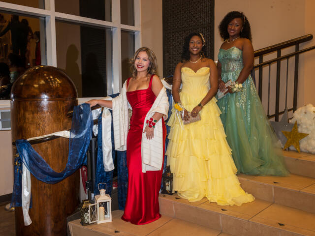 Three women in formal gowns pose on a staircase at a decorated event: red dress, yellow ballgown, and teal gown smiling at the camera.