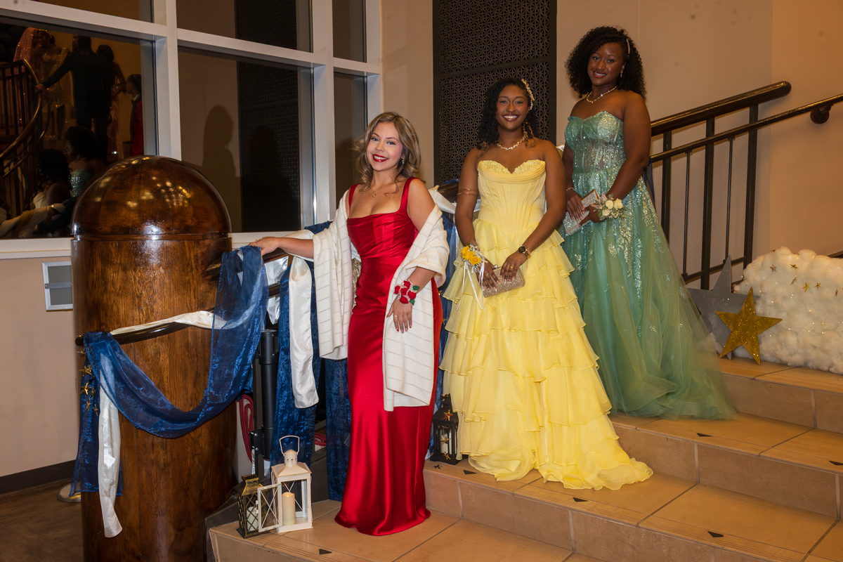 Three women in formal gowns pose on a staircase at a decorated event: red dress, yellow ballgown, and teal gown smiling at the camera.