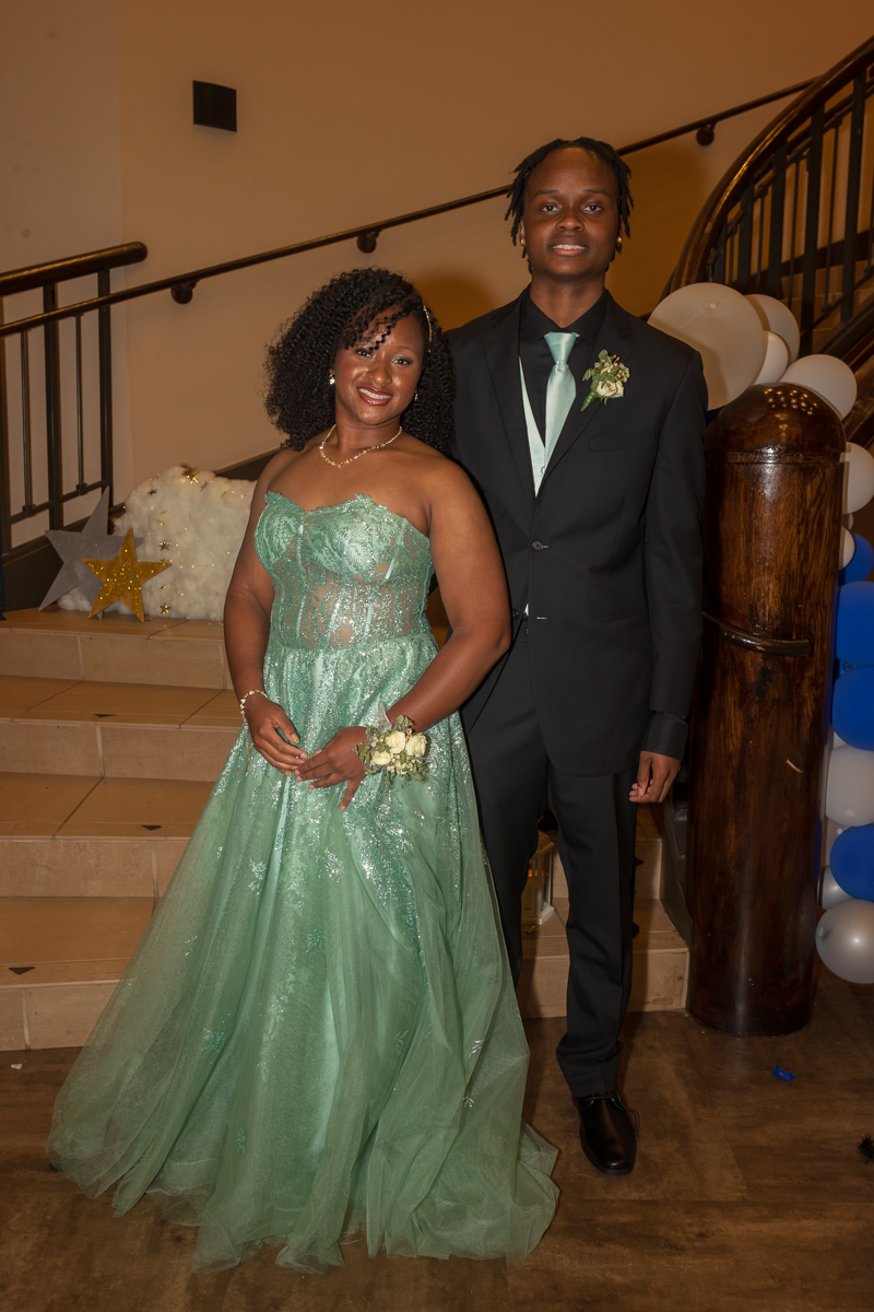 Couple posing on stairs at a formal event: woman in a mint-green gown with a corsage and necklace, man in a black suit and mint tie with a boutonniere.
