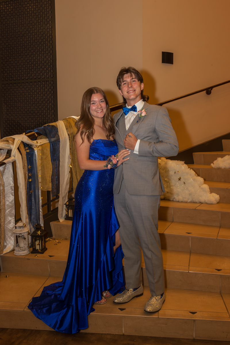 A couple in formal attire posing on a staircase; woman in a blue strapless gown with a corsage and the man in a gray suit with a blue bow tie, smiling at the camera.