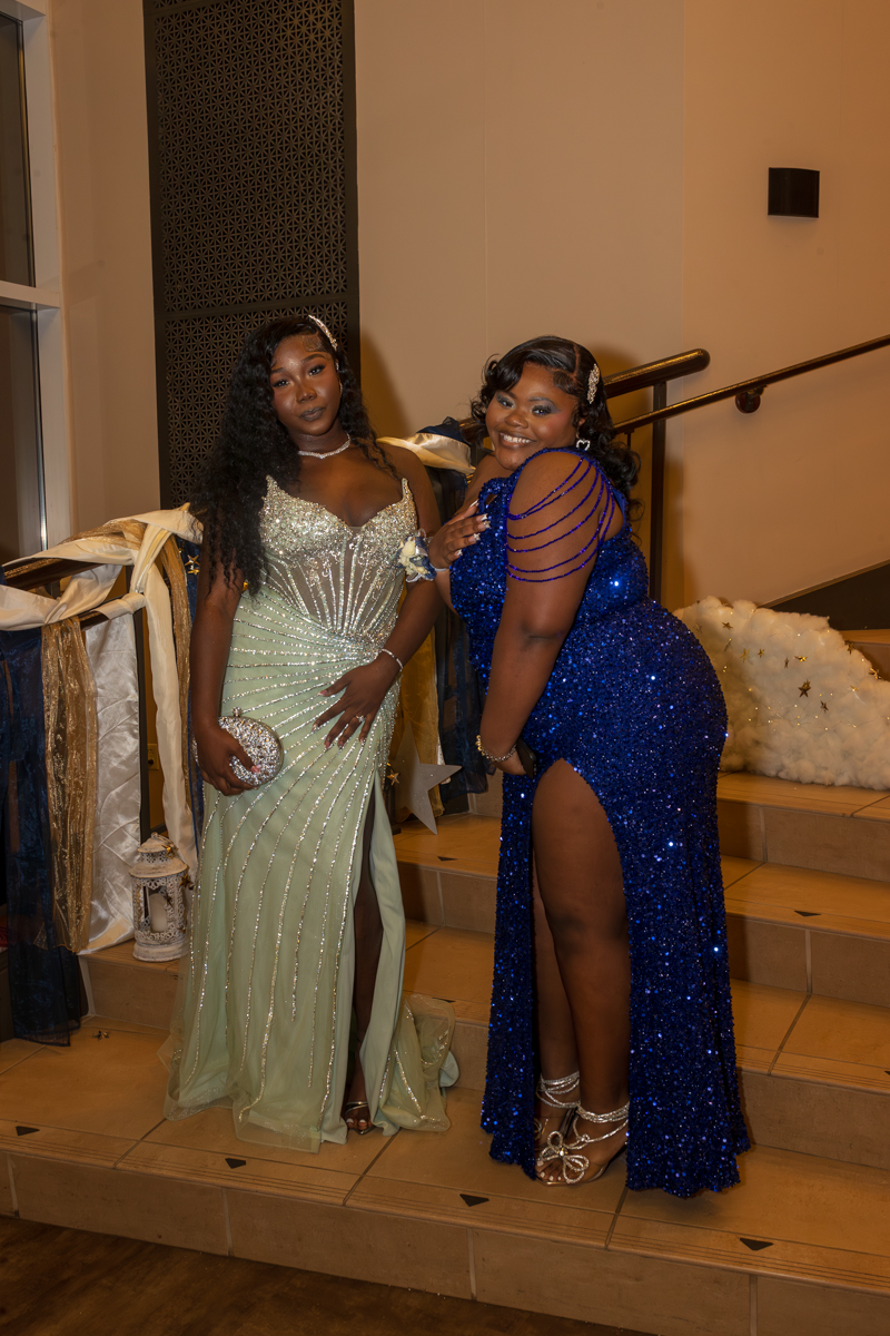 Two women in formal gowns posing on a staircase; one in a pale green beaded gown with a thigh-high slit, the other in a blue sequined gown with a side slit.