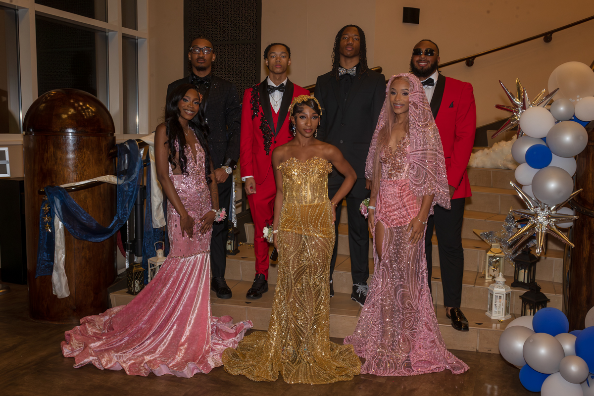 A group of seven stylish young adults in formal gowns and suits posing on a staircase at a gala, with blue, white, and silver balloons and star decor in the background.