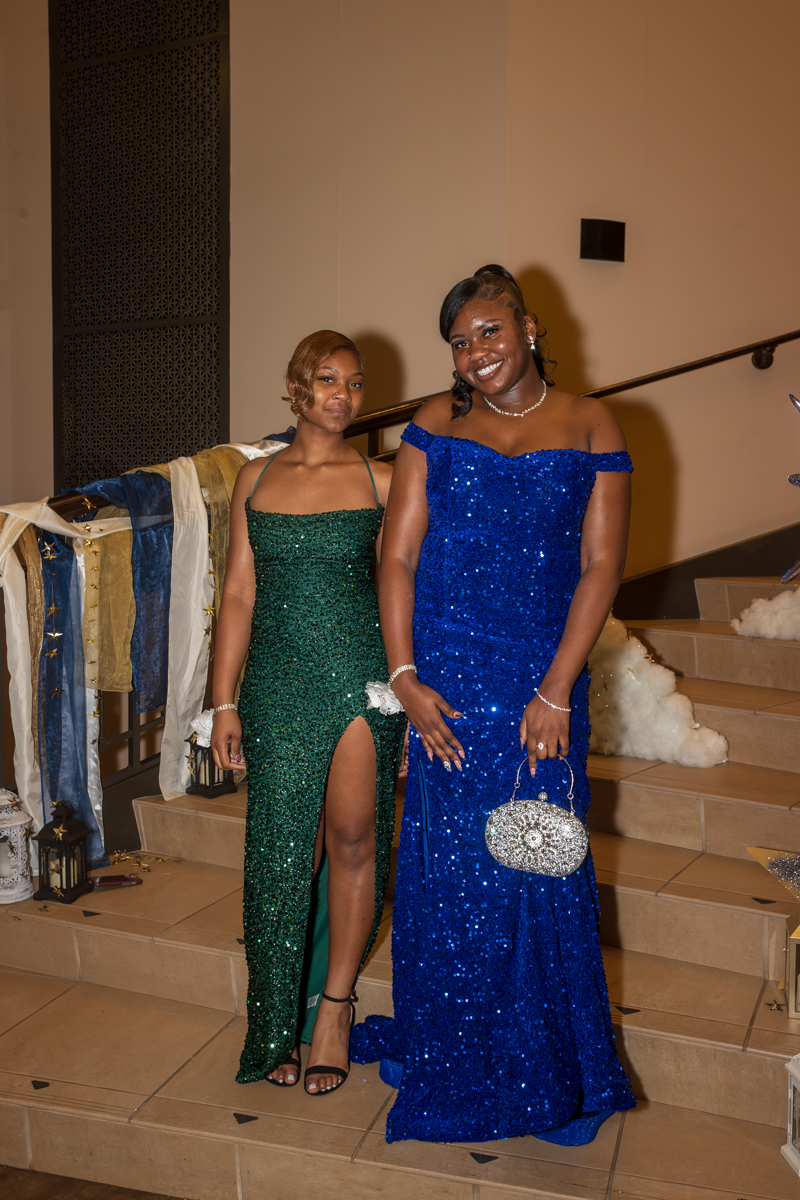 Two women in sequined formal gowns pose on a staircase; one in a green gown with a thigh-high slit and the other in a blue off-shoulder gown with a silver clutch.
