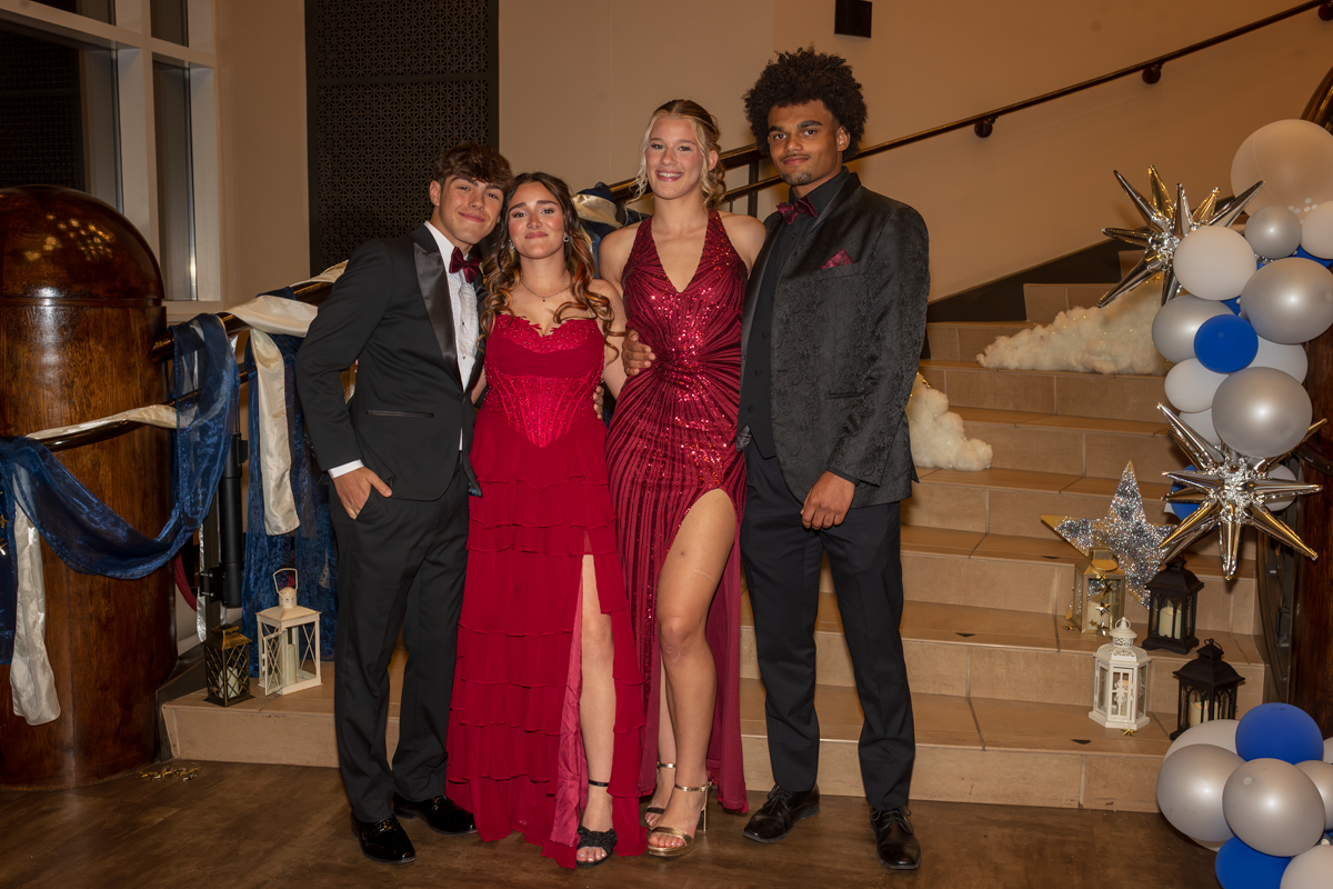 Four friends in formal wear pose on a staircase at a decorated event, two girls in red gowns flanked by two guys in suits.