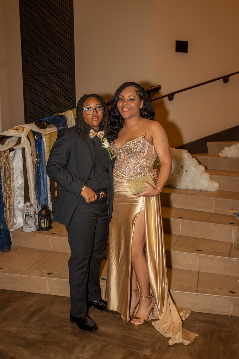 Couple in formal wear posing on a stairway: man in a black tux and bow tie, woman in a gold sequined gown with a thigh-high slit, holding a gold clutch.